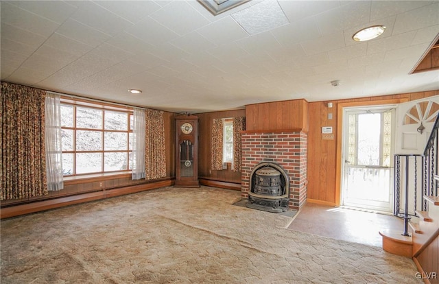 unfurnished living room featuring carpet flooring, wood walls, a wood stove, and a baseboard radiator