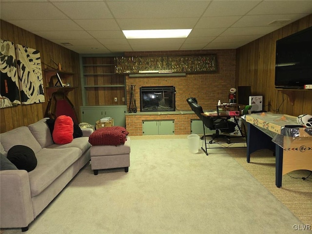 carpeted living room with built in features, wood walls, a brick fireplace, and a drop ceiling