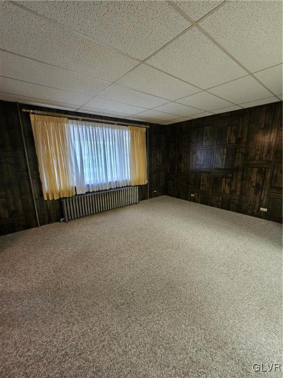 carpeted empty room featuring a drop ceiling, wood walls, and radiator heating unit