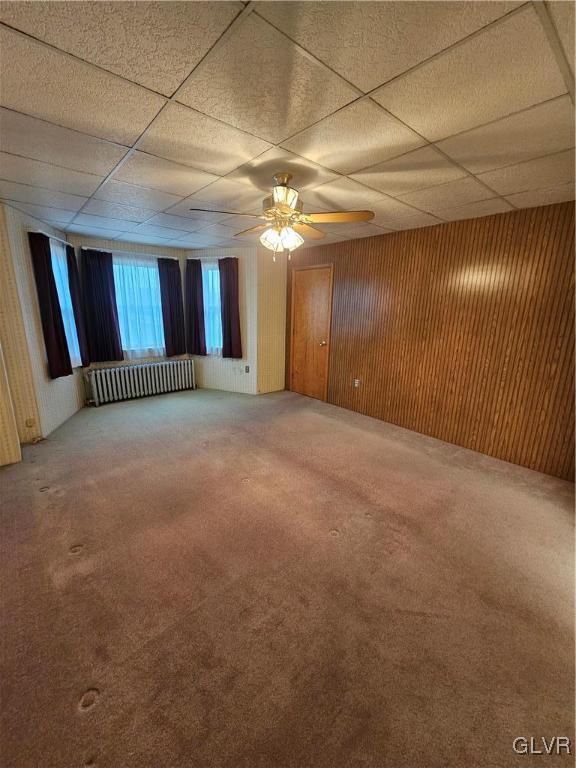 carpeted empty room featuring ceiling fan, a drop ceiling, wood walls, and radiator heating unit