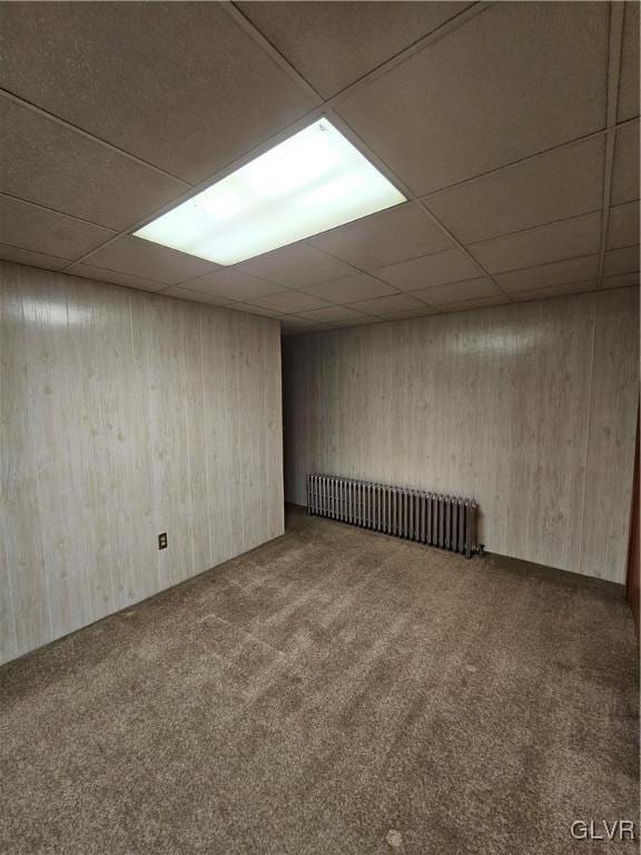 carpeted empty room with radiator, wood walls, and a drop ceiling