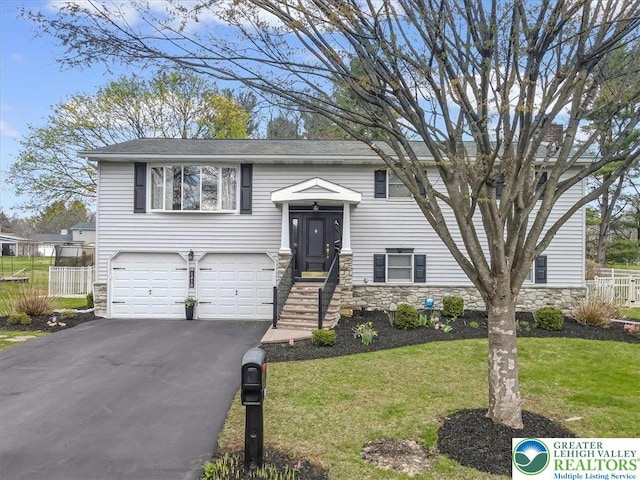 raised ranch featuring driveway, stone siding, a front lawn, a garage, and fence