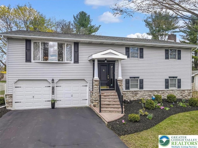 raised ranch with a chimney, stone siding, a garage, and aphalt driveway