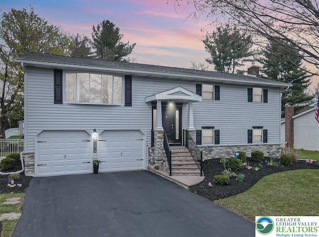raised ranch with a garage, driveway, stone siding, and a chimney