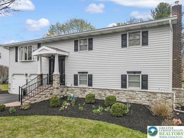 split foyer home featuring driveway, stone siding, a garage, and a chimney