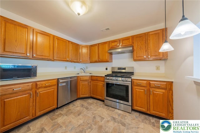 kitchen featuring brown cabinetry, stainless steel appliances, stone finish floor, under cabinet range hood, and a sink