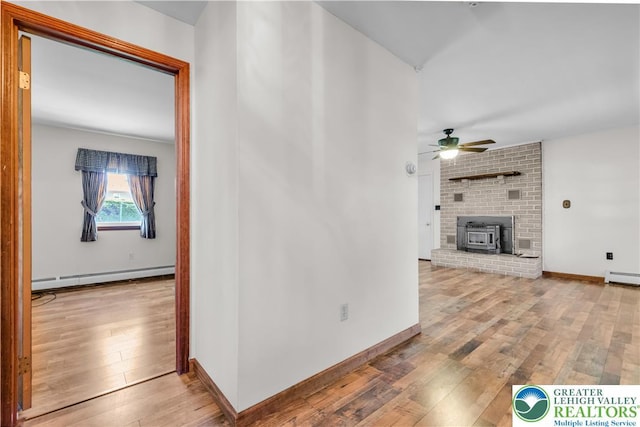 unfurnished living room with hardwood / wood-style flooring, a wood stove, a baseboard radiator, ceiling fan, and baseboards