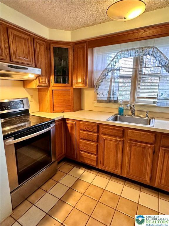 kitchen featuring brown cabinetry, under cabinet range hood, light countertops, a sink, and stainless steel range with electric stovetop