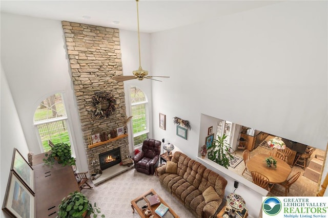 living area with a ceiling fan, a stone fireplace, a healthy amount of sunlight, and a towering ceiling