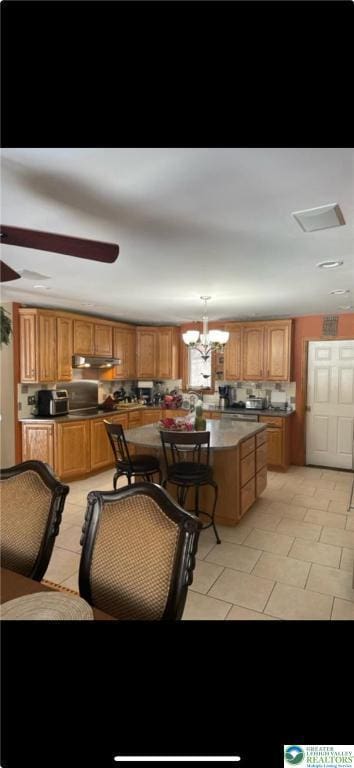 kitchen featuring brown cabinets, ceiling fan with notable chandelier, a center island, light tile patterned floors, and under cabinet range hood