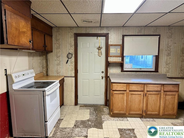 kitchen featuring brown cabinets, white range with electric stovetop, light countertops, wallpapered walls, and a drop ceiling