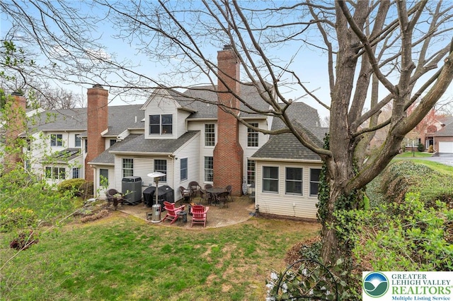 back of house featuring a chimney, roof with shingles, a lawn, central AC unit, and a patio