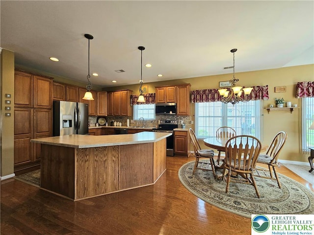 kitchen with brown cabinetry, a kitchen island, appliances with stainless steel finishes, backsplash, and a notable chandelier