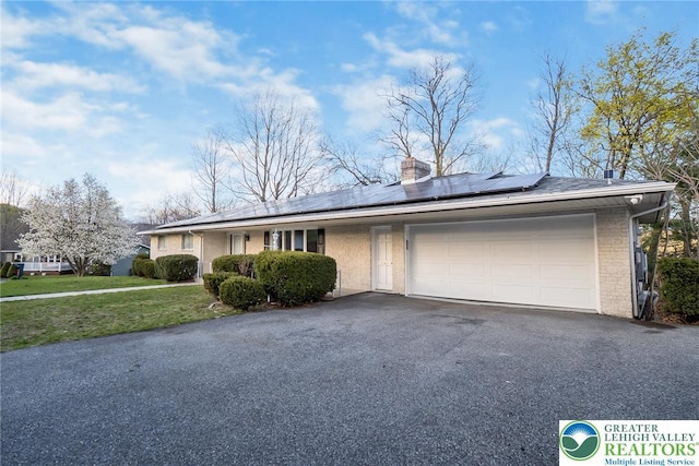 ranch-style home featuring an attached garage, brick siding, aphalt driveway, a chimney, and solar panels