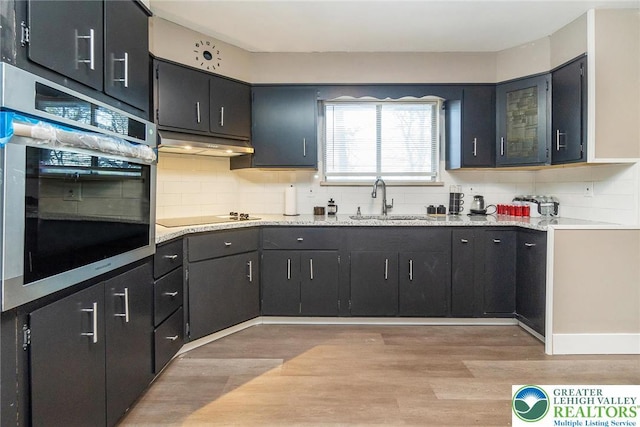 kitchen with oven, black electric stovetop, a sink, light wood-type flooring, and under cabinet range hood