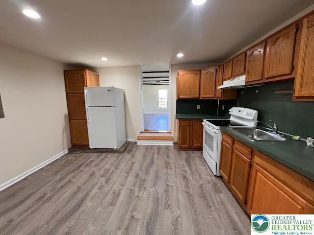 kitchen with dark countertops, a sink, white appliances, brown cabinetry, and light wood-style flooring