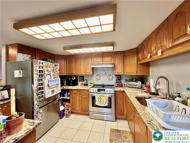 kitchen with brown cabinets, light stone counters, under cabinet range hood, stainless steel appliances, and light tile patterned flooring