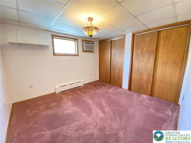 unfurnished bedroom with two closets, a drop ceiling, a wall mounted AC, a baseboard heating unit, and carpet flooring