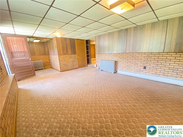 finished basement with carpet, a paneled ceiling, radiator, and wooden walls