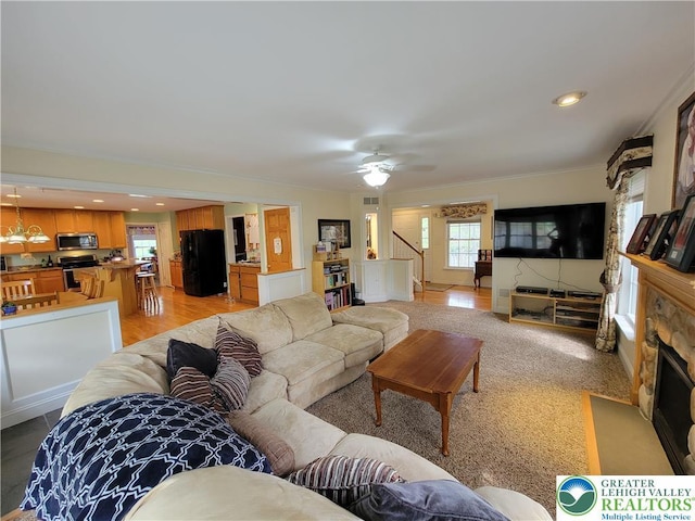 living area with a stone fireplace, ornamental molding, ceiling fan, and stairway