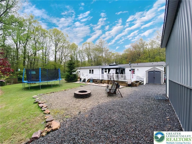 view of grassy yard with an outdoor fire pit, a trampoline, a deck, a storage shed, and a gazebo