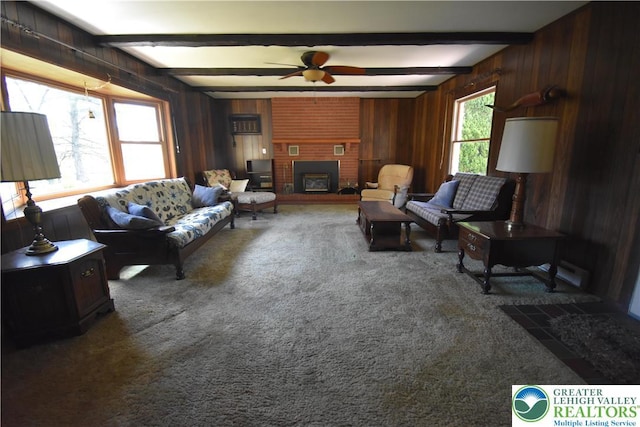 living area featuring carpet flooring, ceiling fan, beamed ceiling, wood walls, and a brick fireplace
