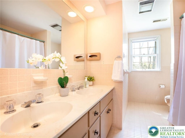 bathroom with tile walls, tile patterned floors, a wainscoted wall, double vanity, and toilet
