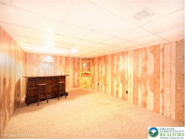 finished basement featuring a bar, carpet, a drop ceiling, and wooden walls