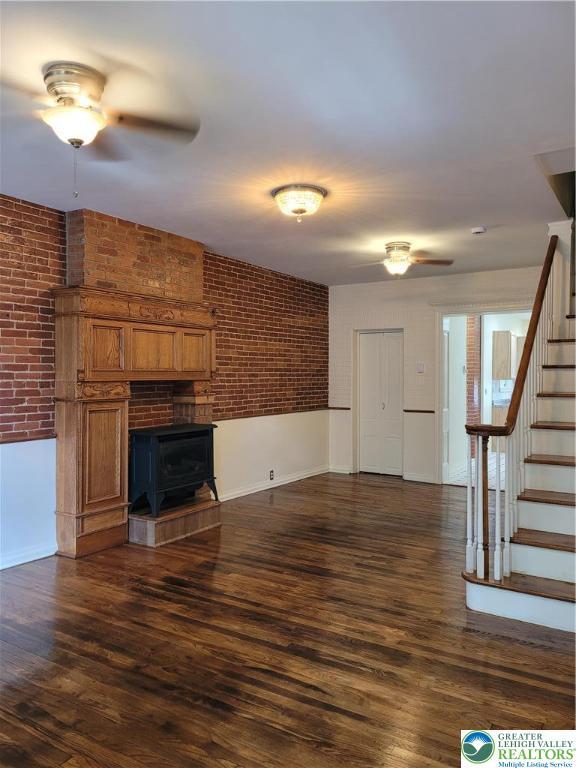 unfurnished living room featuring stairway, a wood stove, brick wall, a ceiling fan, and dark wood-type flooring
