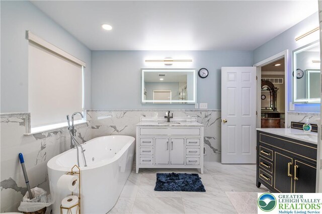 full bath featuring tile walls, wainscoting, a freestanding bath, marble look tile flooring, and two vanities