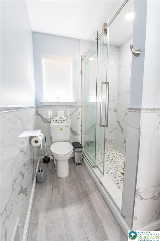 bathroom with tile walls, wood finished floors, a marble finish shower, toilet, and wainscoting