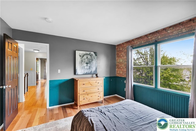 bedroom featuring wainscoting, light wood finished floors, and brick wall