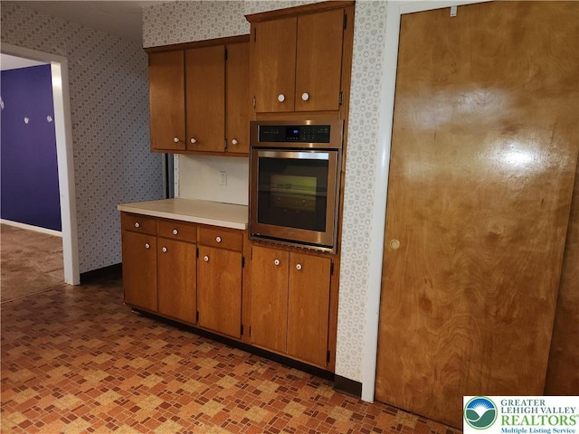 kitchen featuring stainless steel oven, wallpapered walls, light countertops, brown cabinets, and baseboards