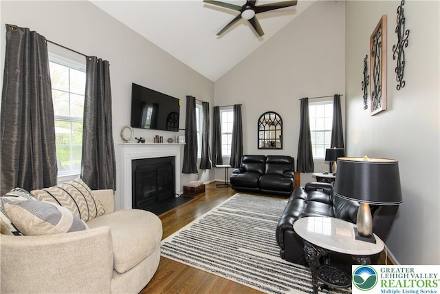 living room with a ceiling fan, wood finished floors, a fireplace with flush hearth, and high vaulted ceiling