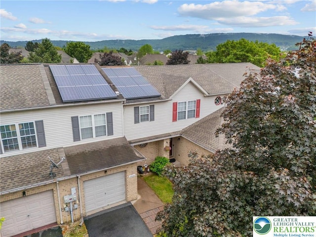 view of property featuring brick siding, driveway, a garage, roof mounted solar panels, and a shingled roof