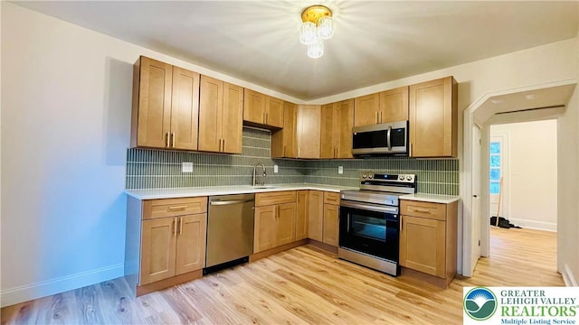 kitchen with appliances with stainless steel finishes, light countertops, a sink, light wood-type flooring, and tasteful backsplash