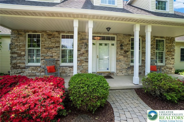doorway to property featuring covered porch, stone siding, and a shingled roof