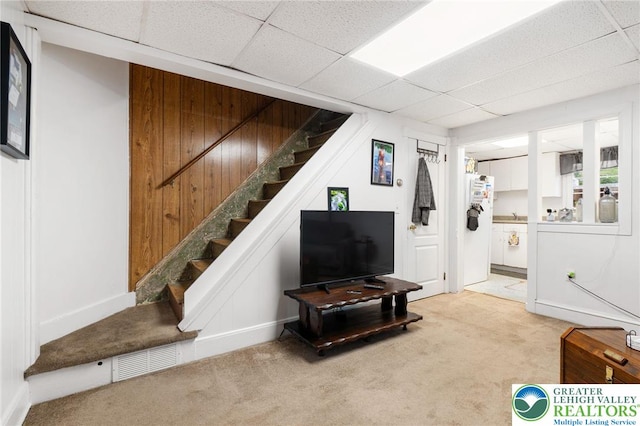 finished below grade area featuring light colored carpet, stairway, a paneled ceiling, freestanding refrigerator, and baseboards