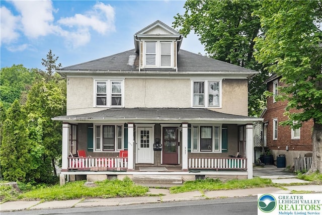 american foursquare style home featuring stucco siding, a porch, roof with shingles, and central AC