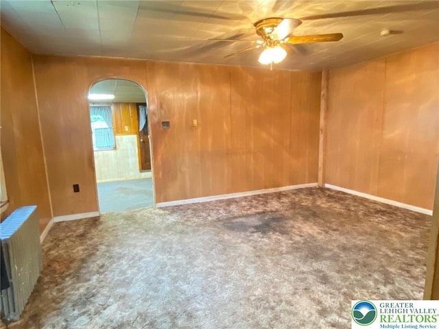 unfurnished room featuring radiator heating unit, arched walkways, ceiling fan, wooden walls, and carpet flooring