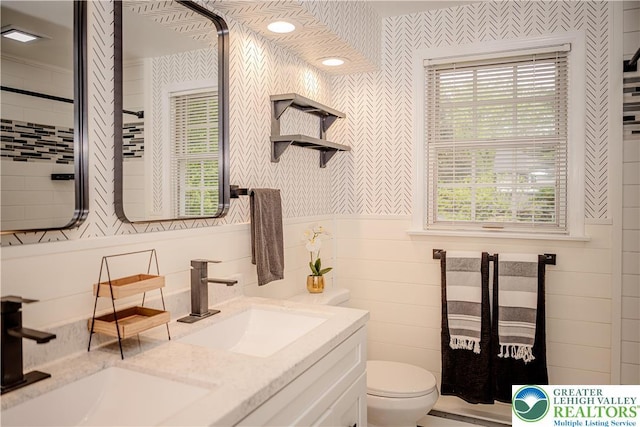 bathroom with wallpapered walls, toilet, double vanity, and wainscoting