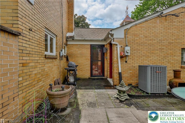 property entrance featuring central AC, brick siding, a shingled roof, and a patio area