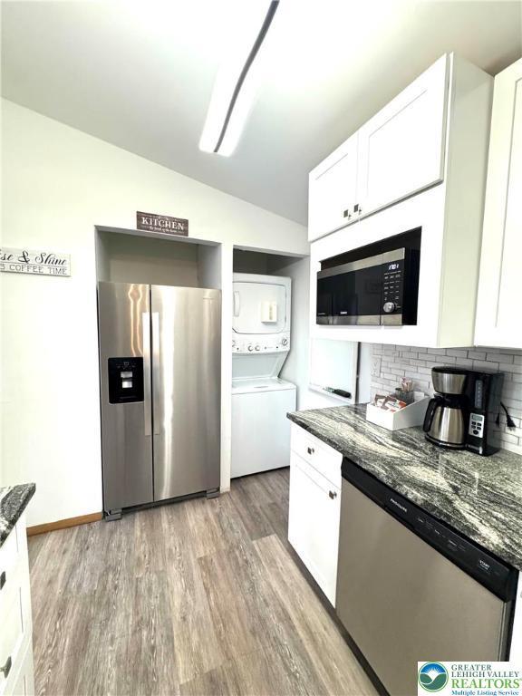 kitchen with appliances with stainless steel finishes, stacked washer and clothes dryer, vaulted ceiling, light wood finished floors, and white cabinetry