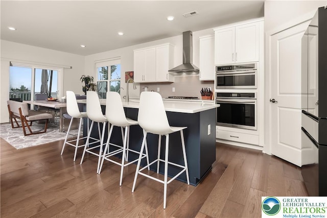 5635 Saucon Ridge Rd, Upper Saucon Twp, PA 18036 - kitchen featuring wall chimney range hood, stainless steel double oven, dark wood-style flooring, a kitchen bar, and a center island with sink