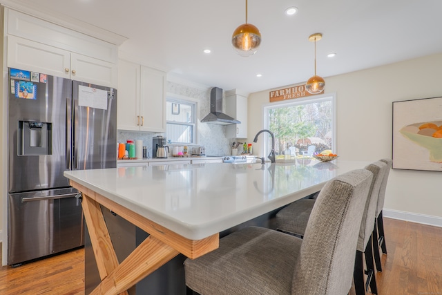 kitchen featuring wall chimney exhaust hood, hanging light fixtures, stainless steel fridge with ice dispenser, light wood-type flooring, and white cabinetry