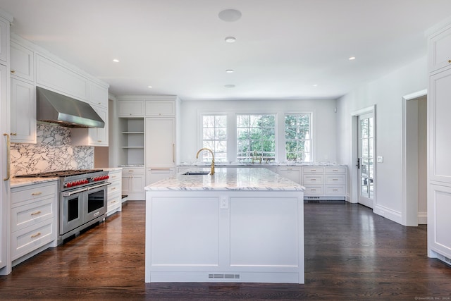 kitchen with white cabinetry, light stone counters, an island with sink, range with two ovens, and wall chimney range hood