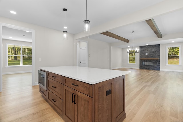 kitchen with brown cabinetry, light wood-style flooring, stainless steel microwave, hanging light fixtures, and beam ceiling