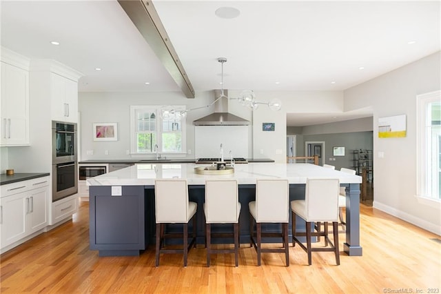 kitchen with wall chimney range hood, light wood-type flooring, appliances with stainless steel finishes, white cabinetry, and a sink