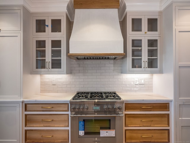 kitchen with stainless steel stove, white cabinets, premium range hood, light stone counters, and backsplash