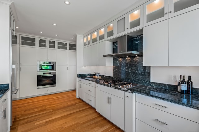 kitchen with tasteful backsplash, white cabinetry, appliances with stainless steel finishes, wall chimney exhaust hood, and light hardwood / wood-style flooring